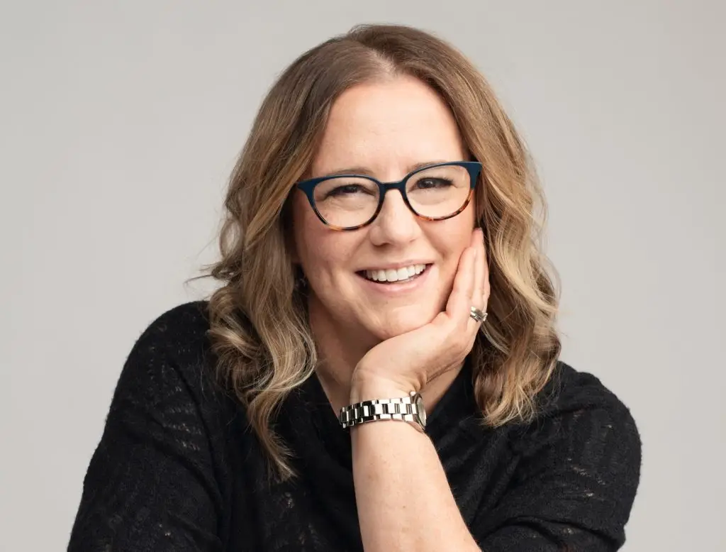 Kim smiles at the camera as she leans forward on a white table, her head resting in her left palm. She has light brown wavy hair that goes past her shoulders. She wears a black quarter-sleave blouse, dark blue glasses and a silver watch.