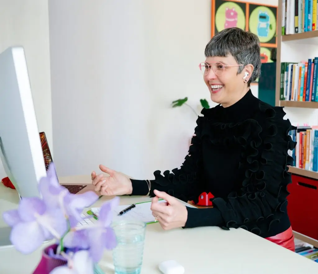 A smiling woman with short grey hair and pink-rimmed glasses sits at a white desk, gesturing with her hands as she looks at a computer monitor.
