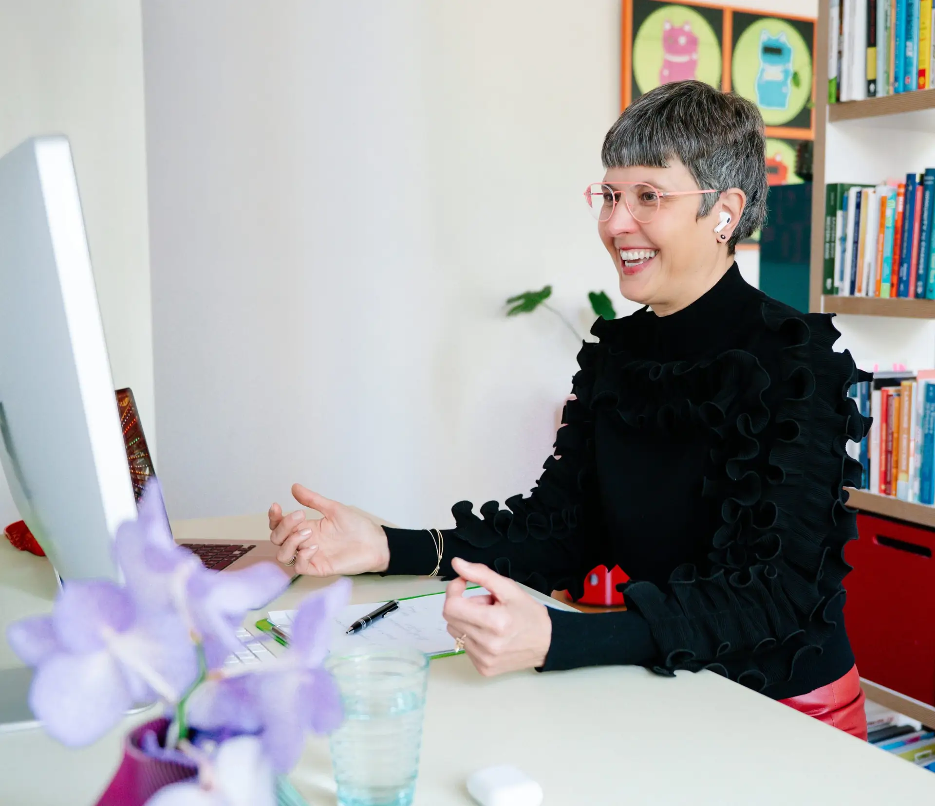 A smiling woman with short grey hair and pink-rimmed glasses sits at a white desk, gesturing with her hands as she looks at a computer monitor.
