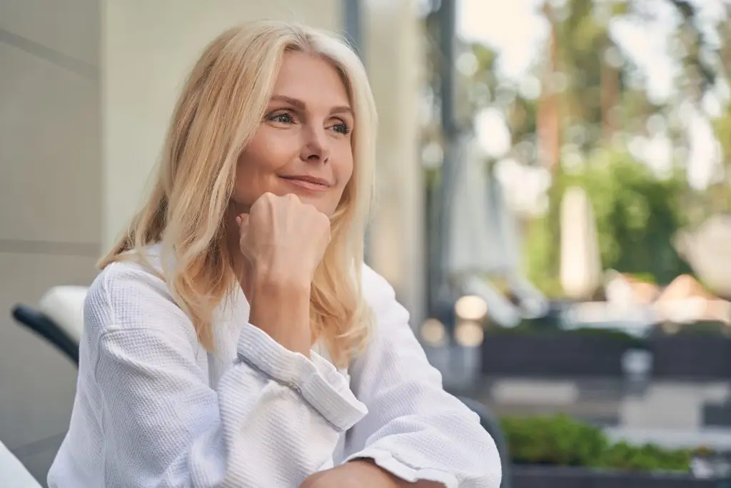 A woman with blonde hair, dressed in a white robe, rests her chin on her fist as she gazes thoughtfully.