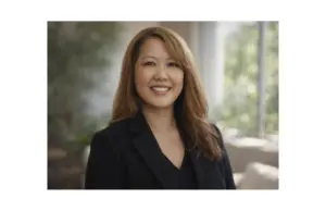 Jacqueline stands facing the camera smiling. She has chest-length light brown hair and wears a v-neck black blouse.