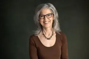 Headshot of Lynda directly facing the camera. She has silver, shoulder length hair. She wears a decroative beaded necklace and a dark brown scoop-necked long-sleaved blouse.