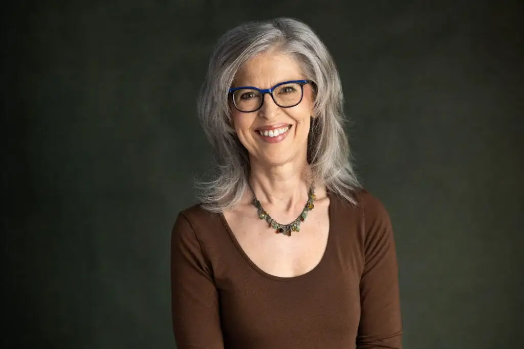 Headshot of Lynda directly facing the camera. She has silver, shoulder length hair. She wears a decroative beaded necklace and a dark brown scoop-necked long-sleaved blouse.