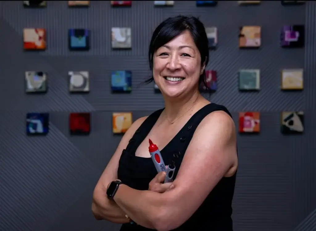 Summer smiles at the camera with confident crossed arms. She waers a black tank top and is holding two bold markers in one hand. A grid of small framed artwork hands behind her on a dark colored wall.
