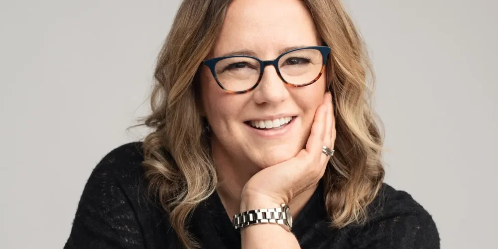 Kim smiles at the camera as she leans forward on a white table, her head resting in her left palm. She has light brown wavy hair that goes past her shoulders. She wears a black quarter-sleave blouse, dark blue glasses and a silver watch.
