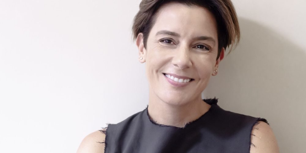 Headshot of Ghada smiling at the camera. She has short, straight, dark brown hair that reaches to her ears. She stands against a white wall wearing a black sleeveless shirt.