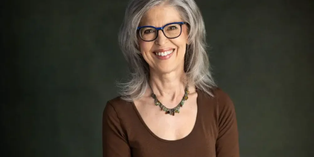 Headshot of Lynda directly facing the camera. She has silver, shoulder length hair. She wears a decroative beaded necklace and a dark brown scoop-necked long-sleaved blouse.