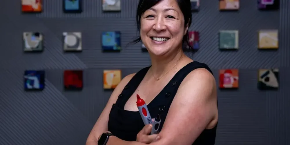 Summer smiles at the camera with confident crossed arms. She waers a black tank top and is holding two bold markers in one hand. A grid of small framed artwork hands behind her on a dark colored wall.
